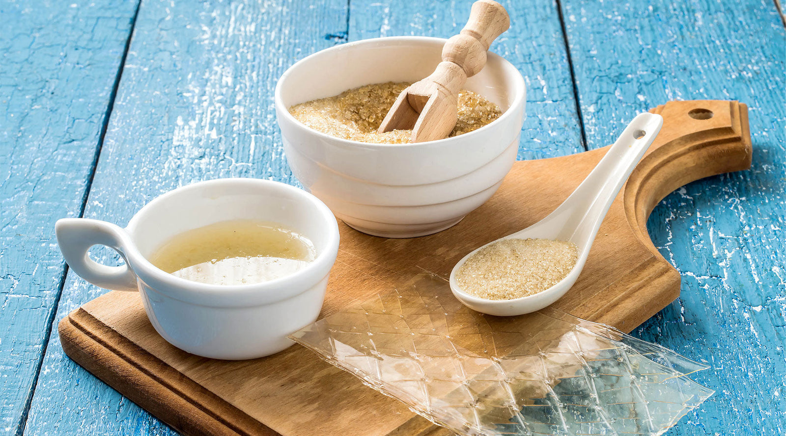White bowls and spoon with a wooden scoop with gelatine powder and leaf gelatine in, on a wooden cutting board against a blue wooden background from MM Ingredients website