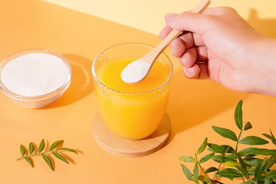 A glass of orange juice with a spoon containing collagen powder just above the glass